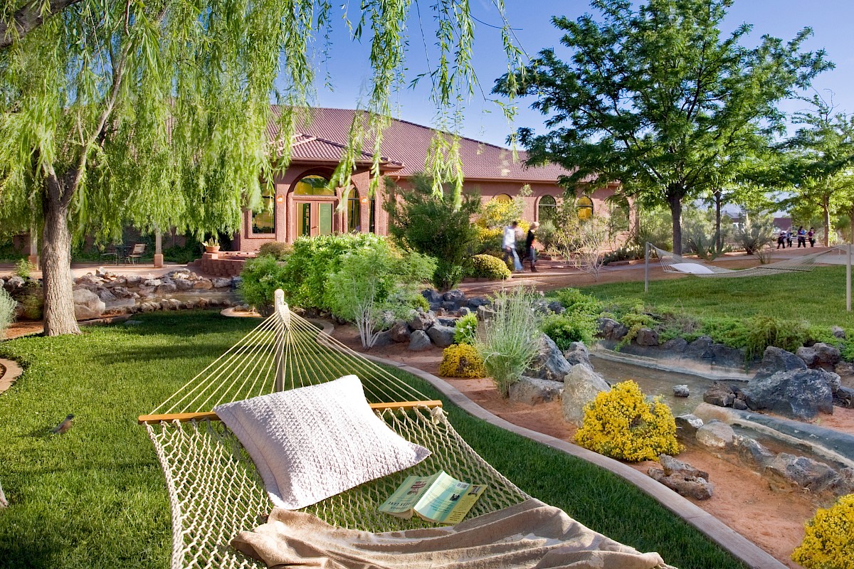A serene garden features a hammock with a pillow, book, and beautiful landscaping, with a house in the background and vibrant greenery ending the sentence.