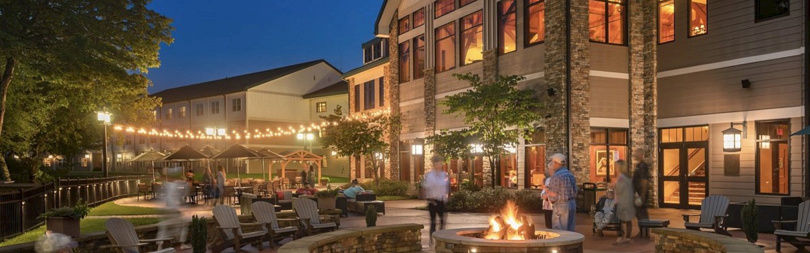 An evening scene of a cozy, spacious patio at a resort with a fire pit, Adirondack chairs, string lights, and several people enjoying the ambiance.
