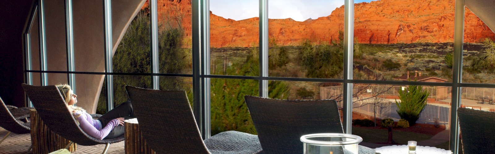 A person relaxes in a modern room with floor-to-ceiling windows offering a stunning view of red rock formations and desert landscape.