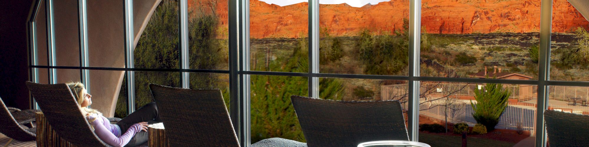 A person relaxes in a modern room with floor-to-ceiling windows offering a stunning view of red rock formations and desert landscape.