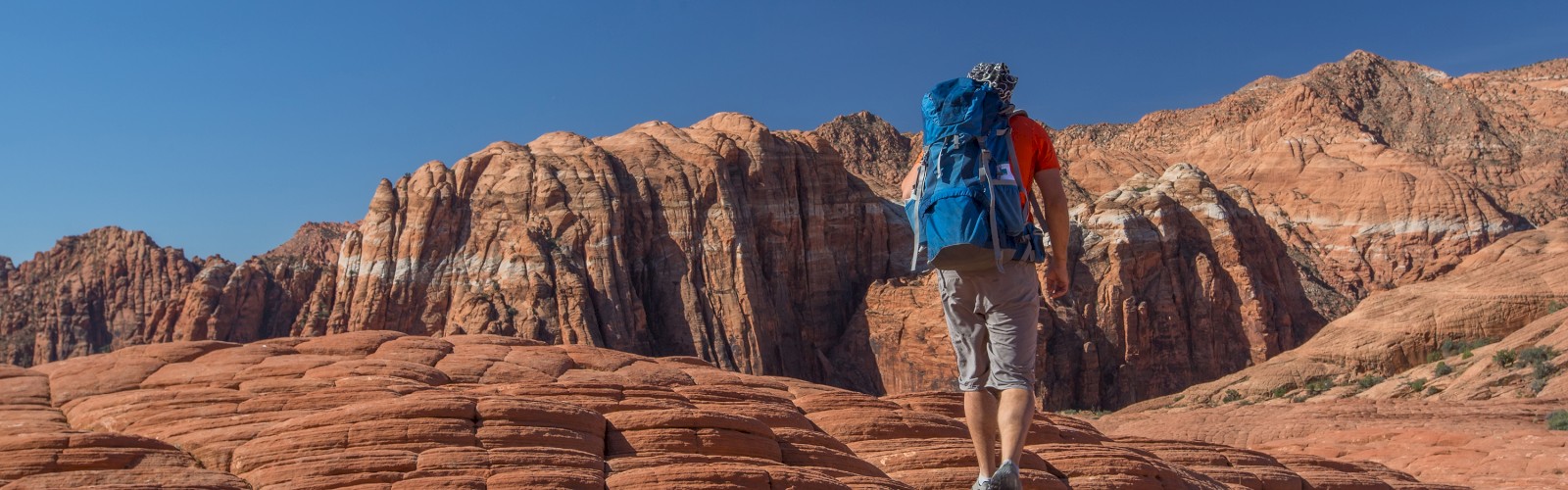 A person with a blue backpack hikes over red rock formations under a clear blue sky in a mountainous desert landscape.