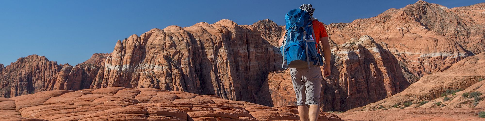 A person with a blue backpack hikes over red rock formations under a clear blue sky in a mountainous desert landscape.