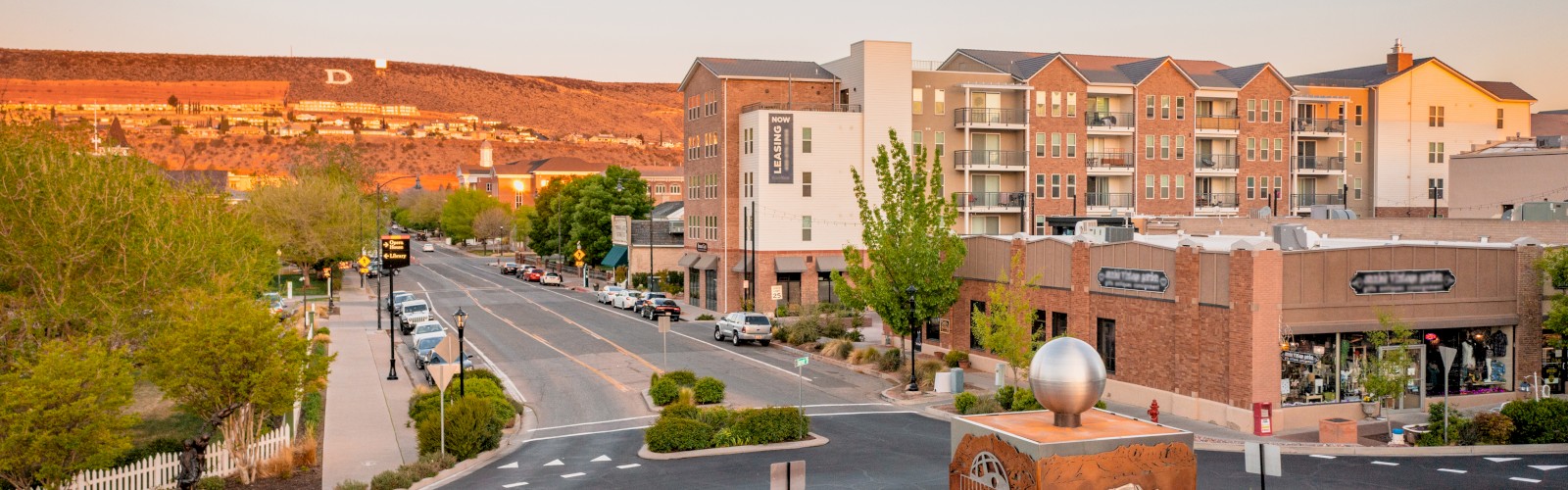 Aerial view of a small town intersection with a roundabout, surrounded by buildings, shops, trees, and mountains in the background, ending the sentence.