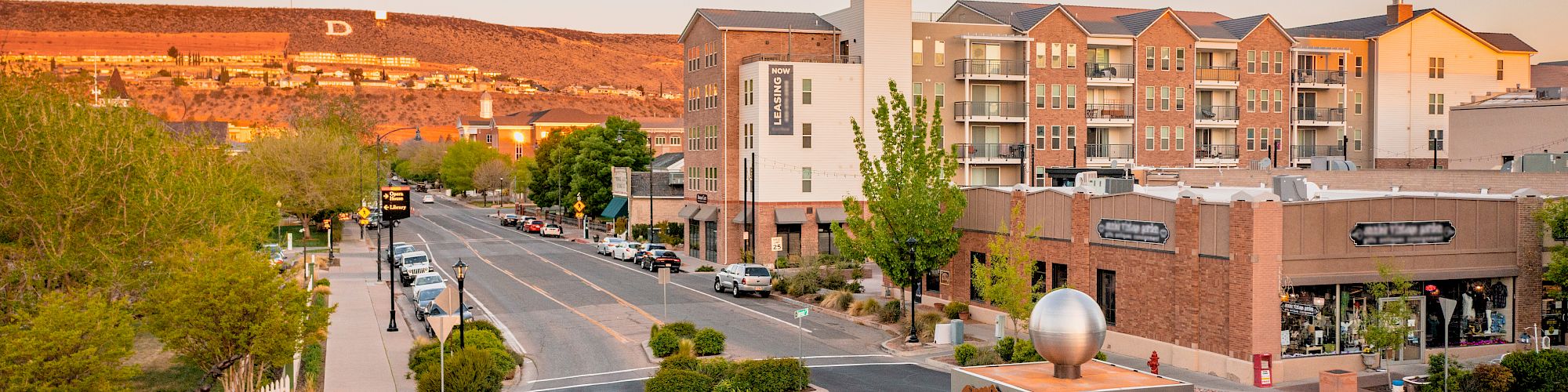 Aerial view of a small town intersection with a roundabout, surrounded by buildings, shops, trees, and mountains in the background, ending the sentence.