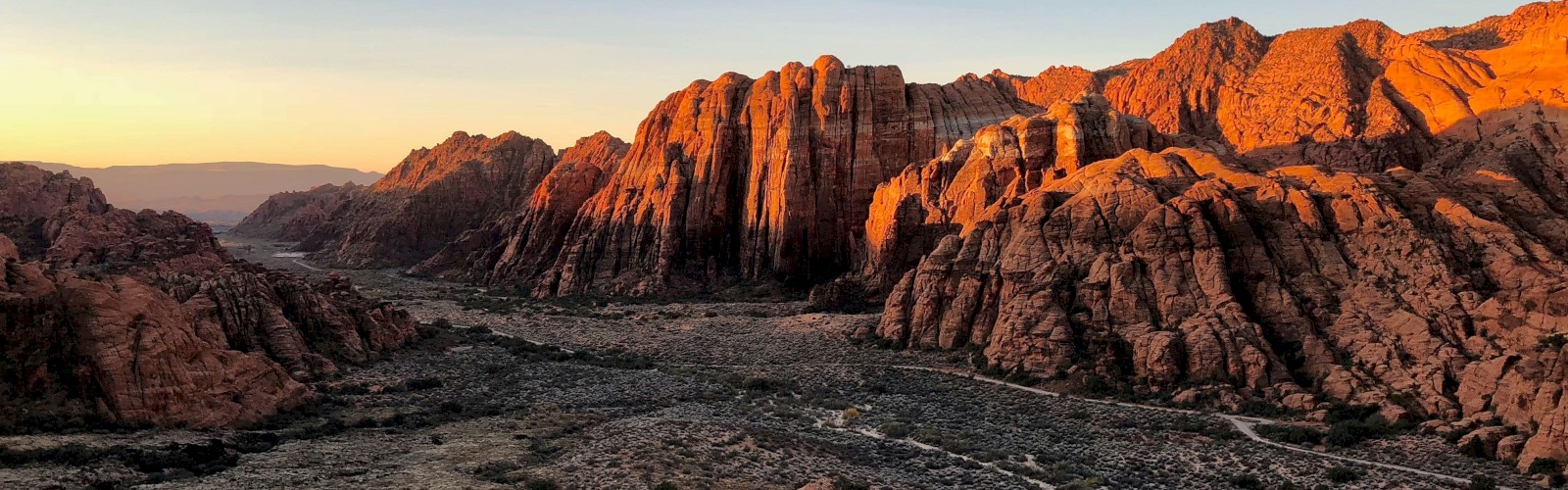 A beautiful landscape of rugged, sunlit red rock formations at sunset, with a dry, rocky valley below and a clear, expansive sky above.