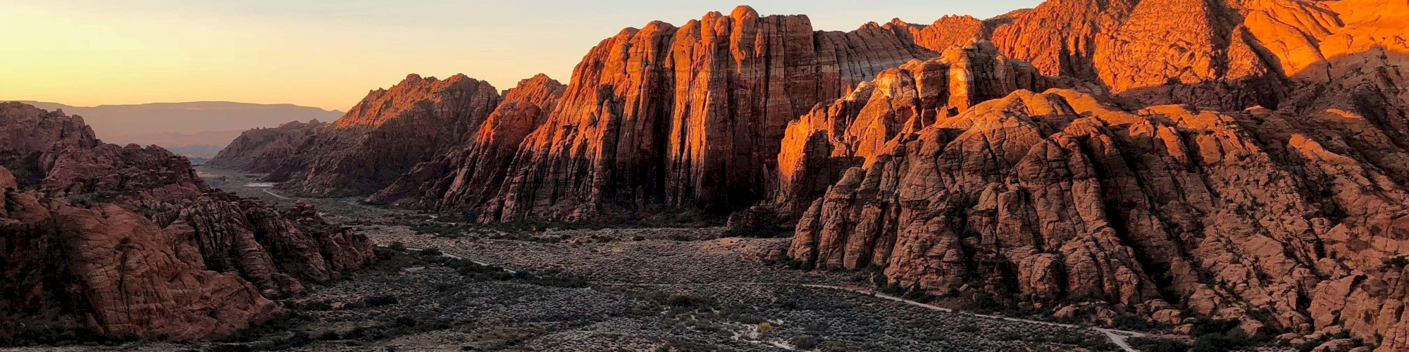 A beautiful landscape of rugged, sunlit red rock formations at sunset, with a dry, rocky valley below and a clear, expansive sky above.