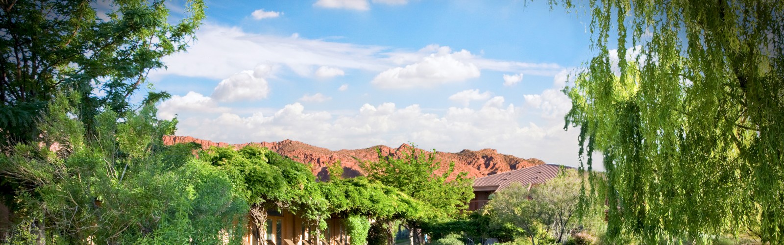 A serene garden features a flowing stream, lush greenery, trees, and manicured lawns under a bright blue sky with distant red mountains ending the sentence.