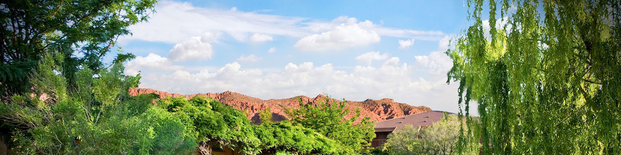 A serene garden features a flowing stream, lush greenery, trees, and manicured lawns under a bright blue sky with distant red mountains ending the sentence.