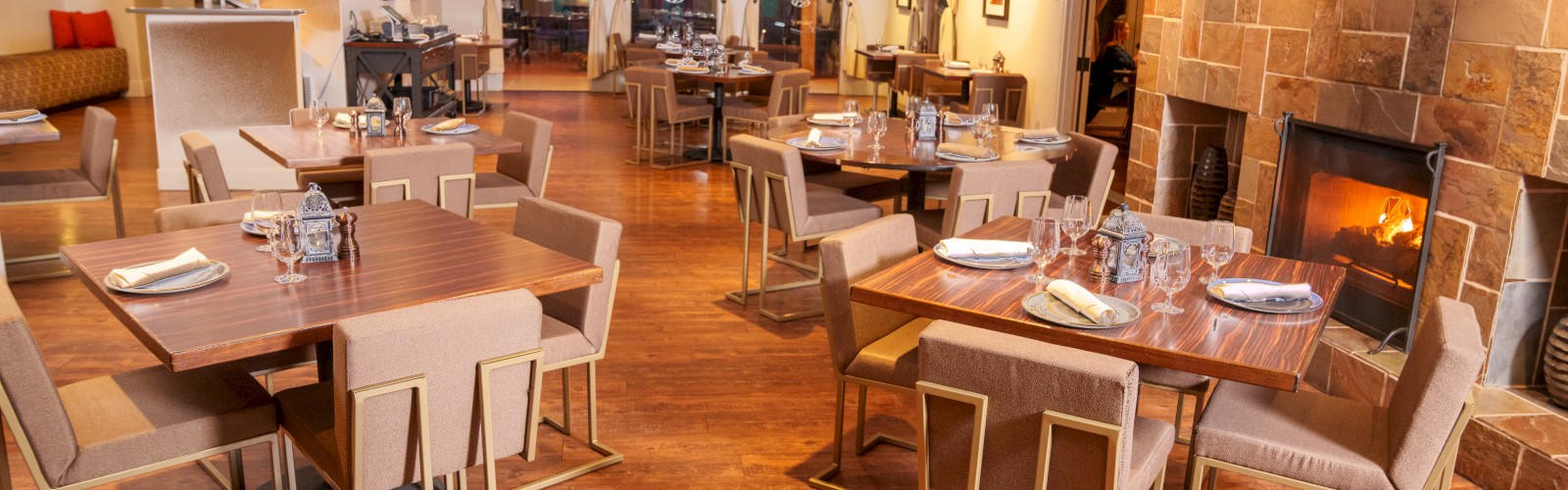 A cozy restaurant interior with wooden flooring, modern tables and chairs, a fireplace, and dim lighting for a warm ambiance, ending the sentence.