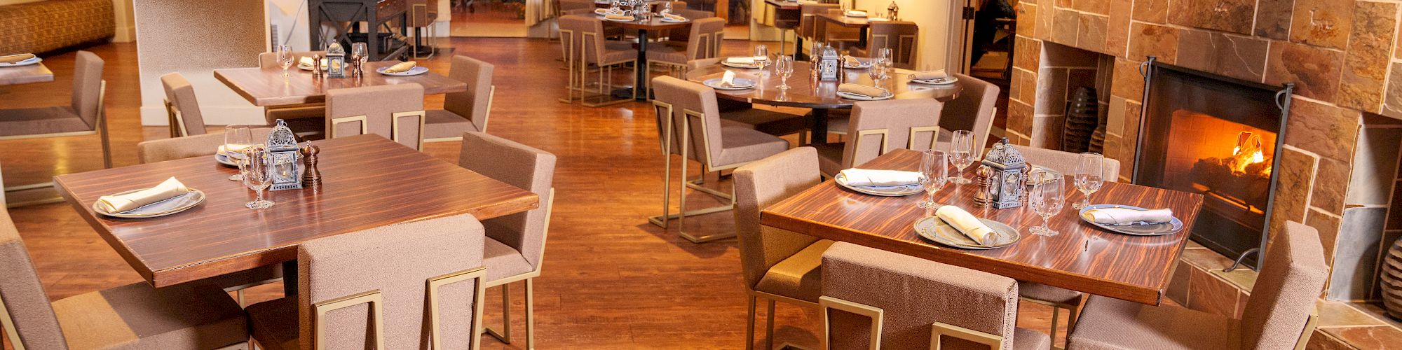 A cozy restaurant interior with wooden flooring, modern tables and chairs, a fireplace, and dim lighting for a warm ambiance, ending the sentence.