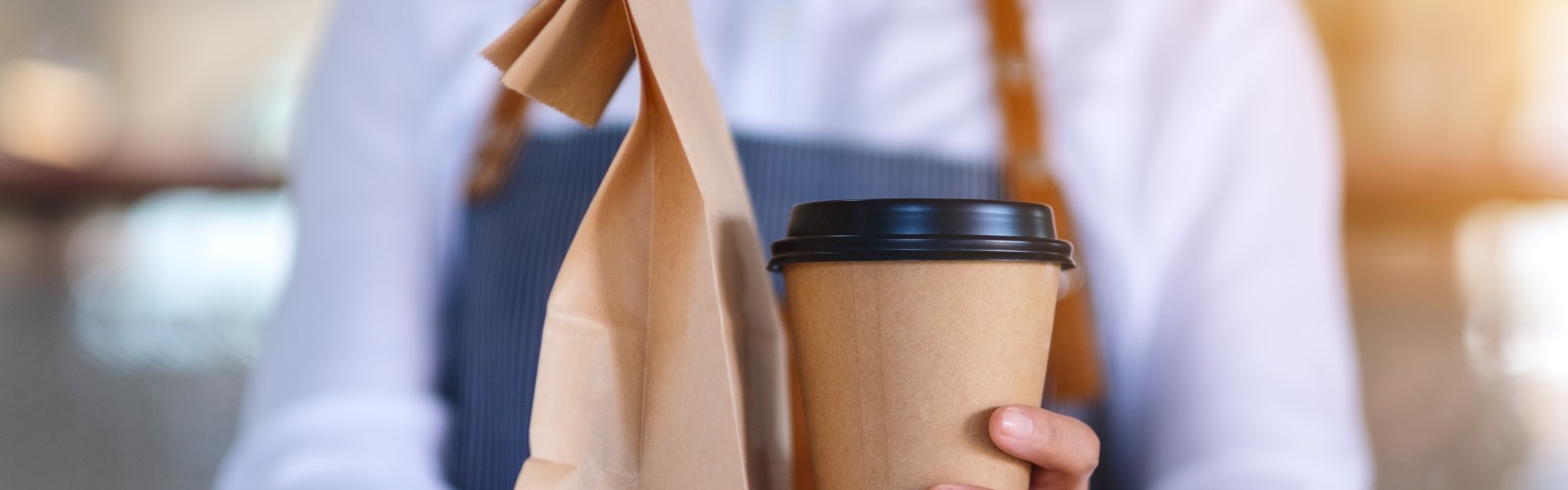 A person in an apron is holding a paper bag and a takeaway coffee cup, suggesting they are serving or delivering food and drink.