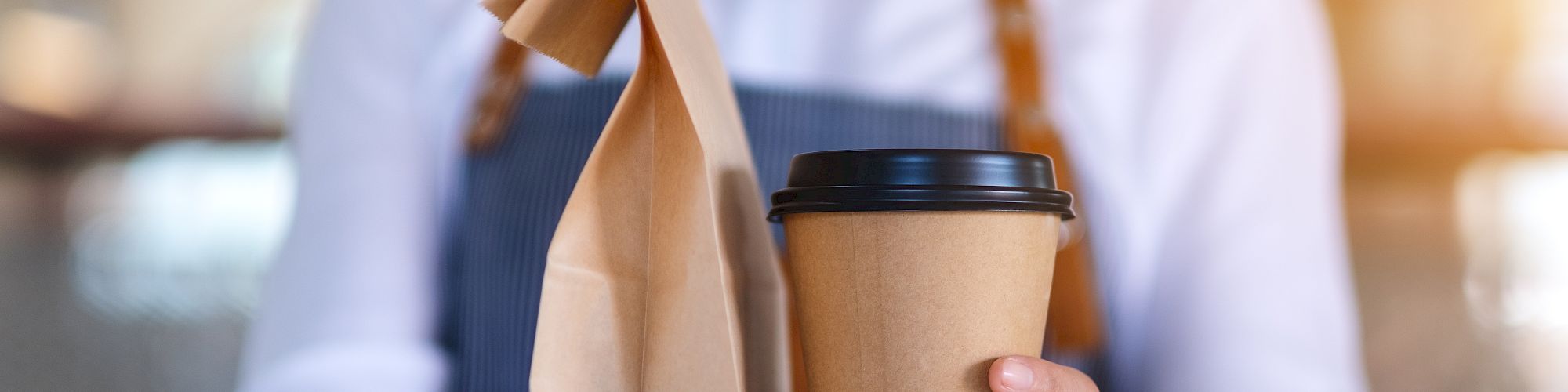 A person in an apron is holding a paper bag and a takeaway coffee cup, suggesting they are serving or delivering food and drink.
