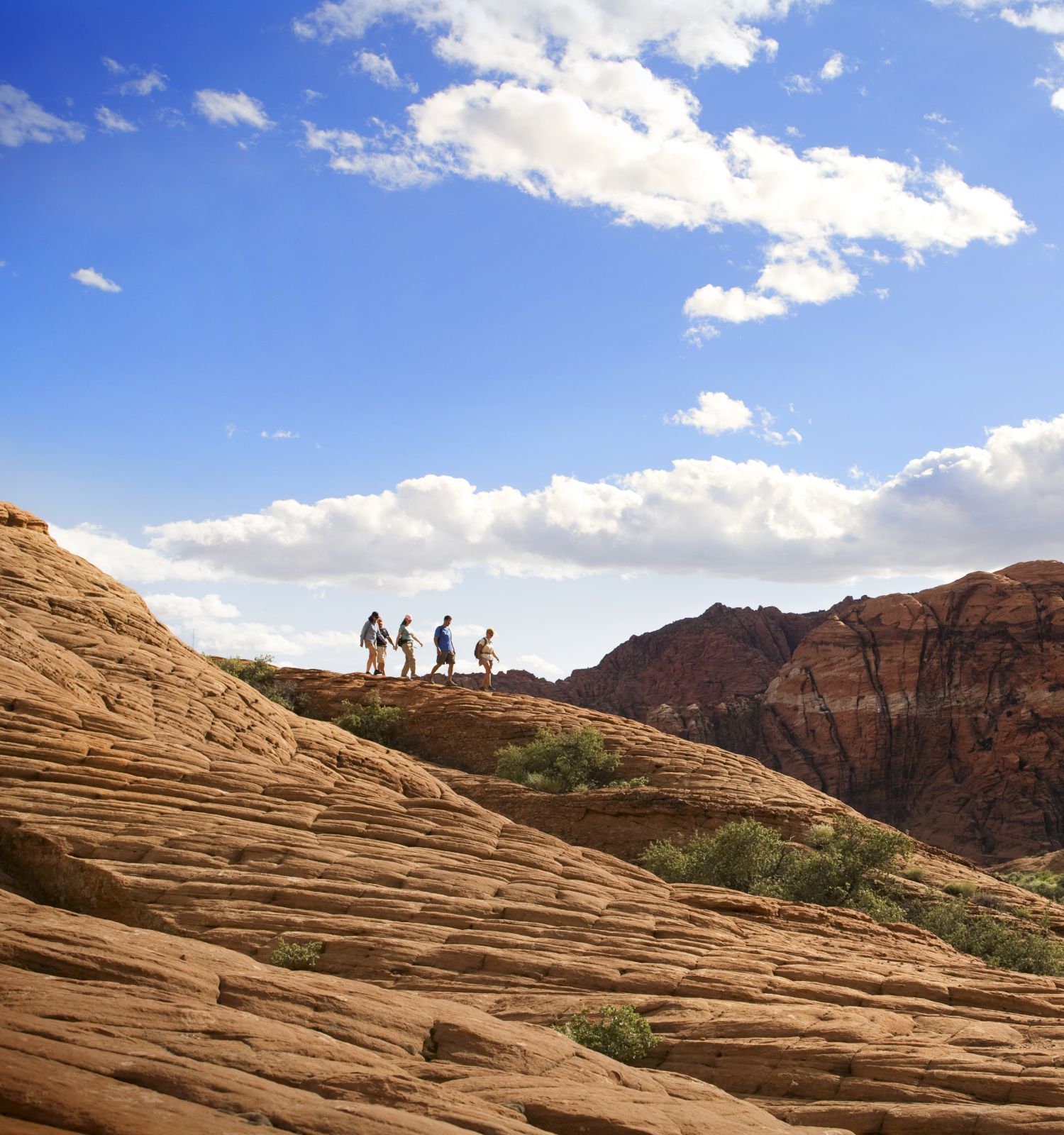 Four people walk along the top of a rocky hill with a mountainous background and a blue sky with scattered clouds.