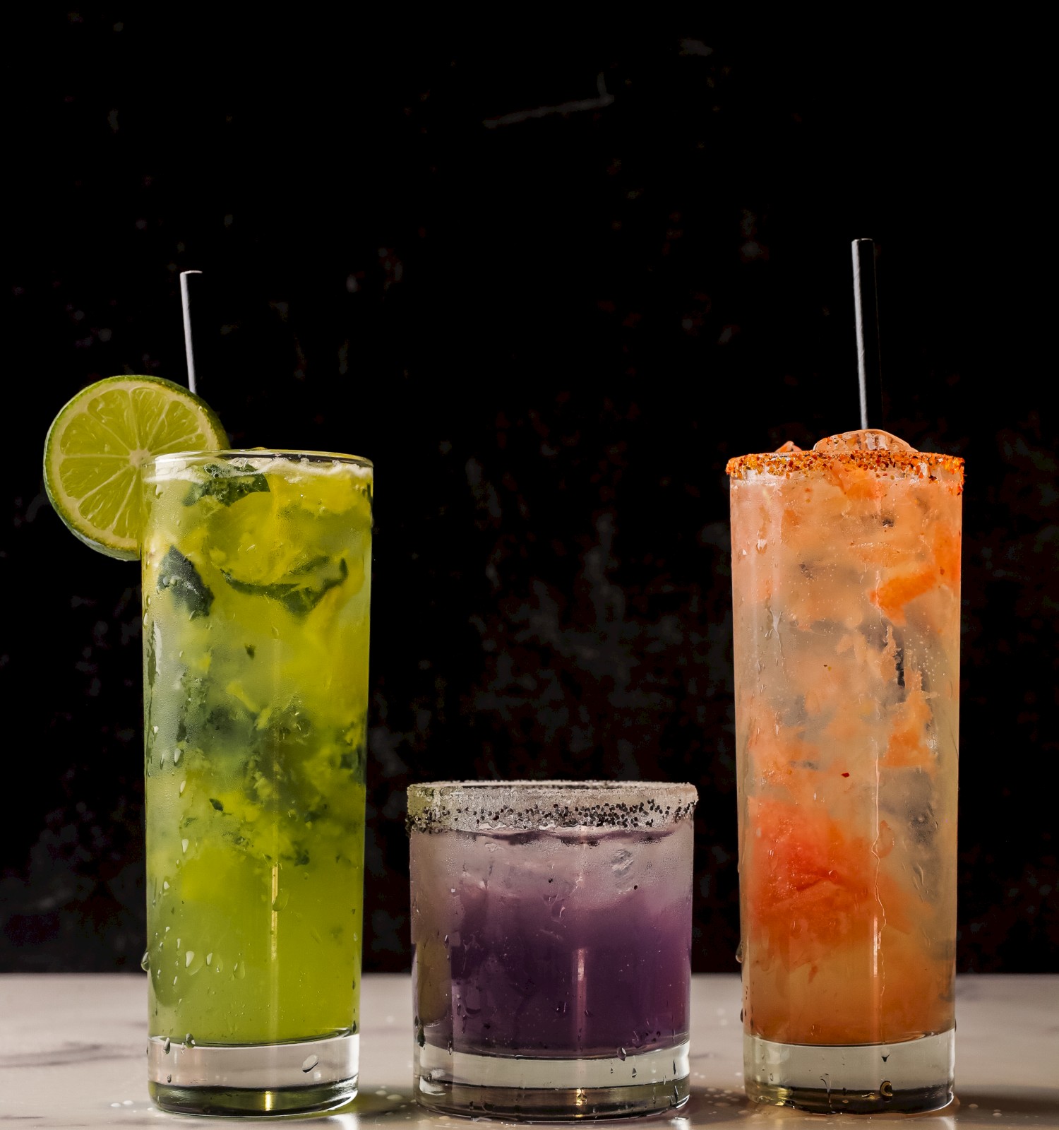 Three colorful cocktails: a green drink with a lime slice, a purple drink, and an orange drink with ice. They are placed on a white surface.