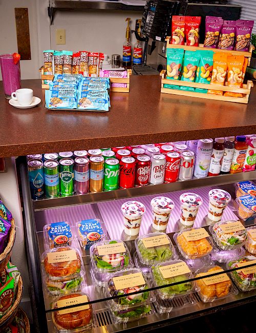 The image shows a store counter with snacks, beverages, and salads on display. A basket has packaged snacks; an assortment of colorful drinks is visible.