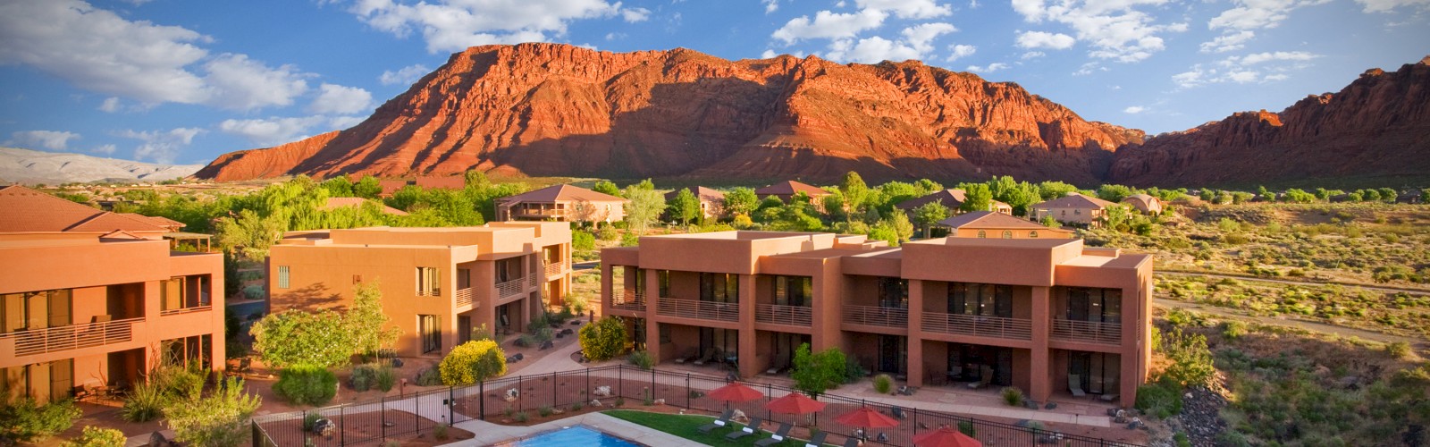 A resort with a swimming pool, sun loungers, and red umbrellas, set against a backdrop of red rock formations and a bright, partly cloudy sky.