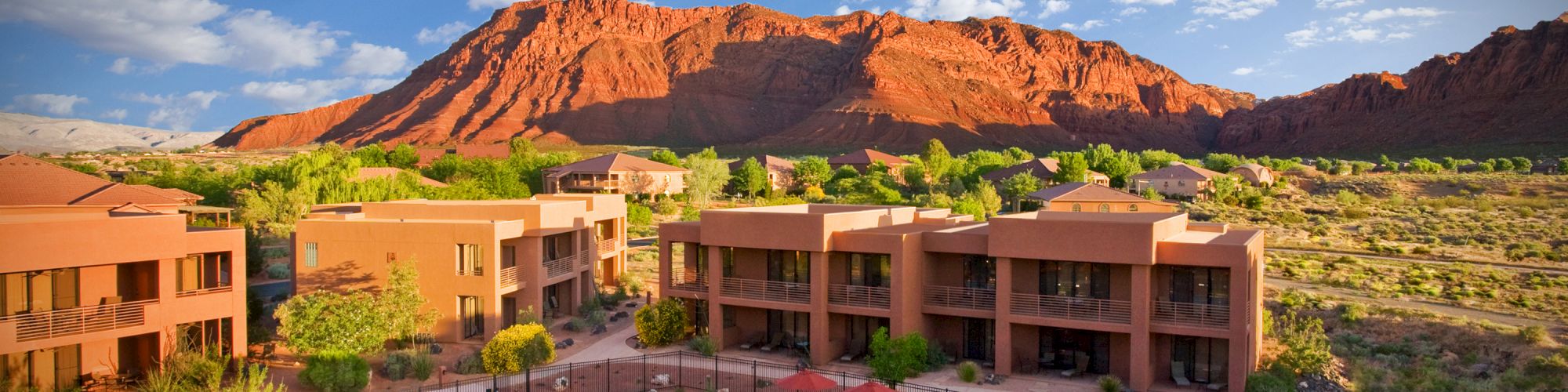 A resort with a swimming pool, sun loungers, and red umbrellas, set against a backdrop of red rock formations and a bright, partly cloudy sky.