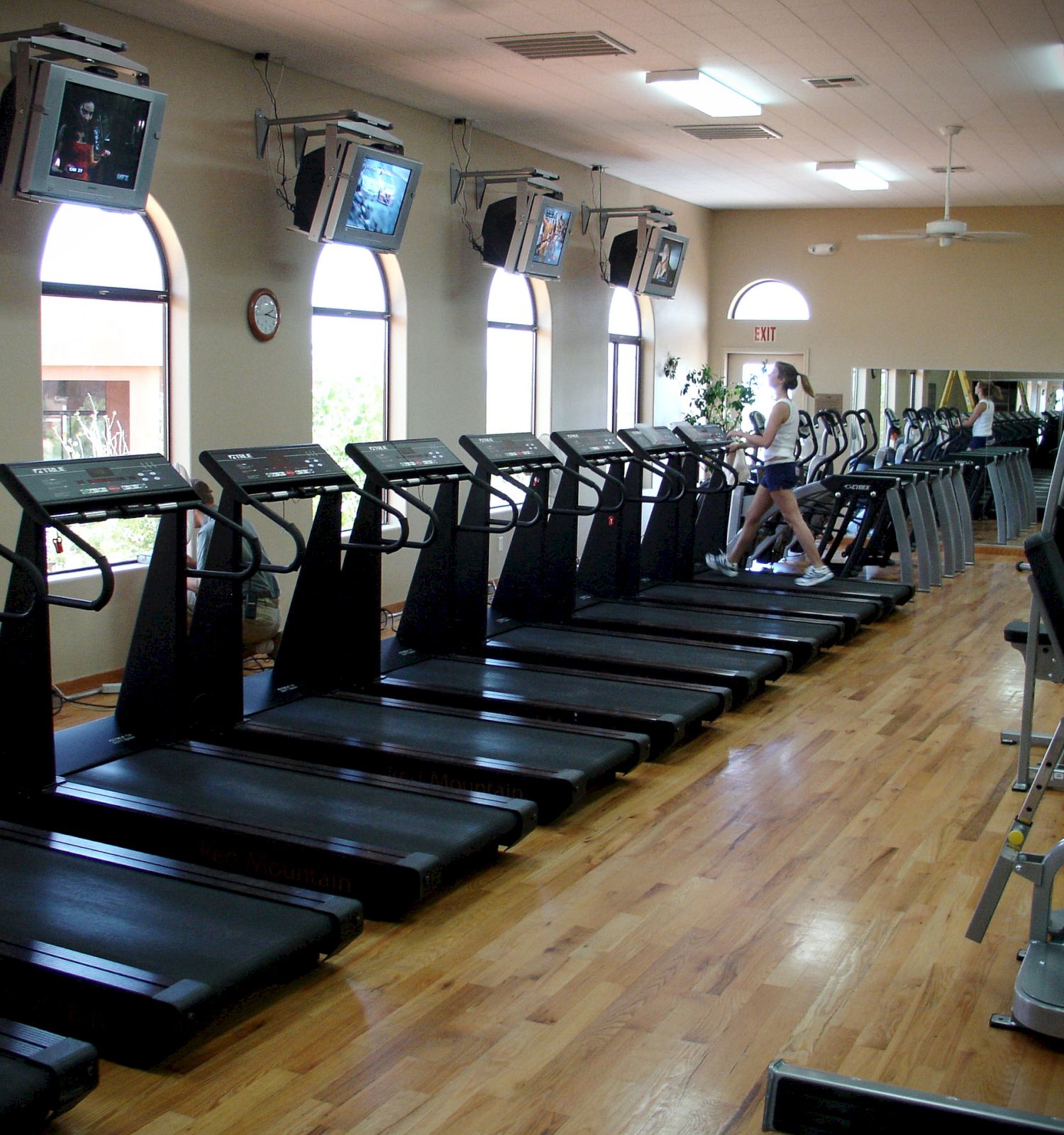 A gym with multiple treadmills lined up, a person exercising, wall-mounted TVs, large windows, and wooden flooring. Equipment seen in the foreground.