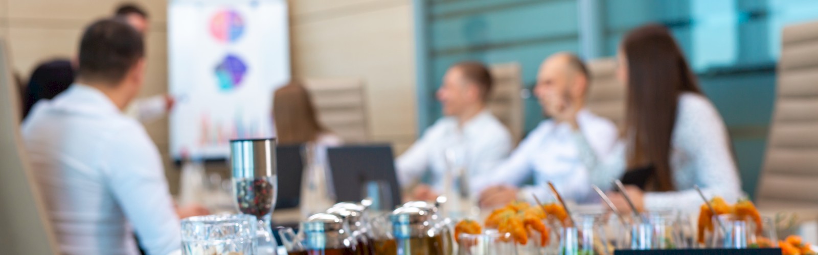 A business meeting is taking place with a diverse array of appetizers and drinks on the table, while a person points at a whiteboard.