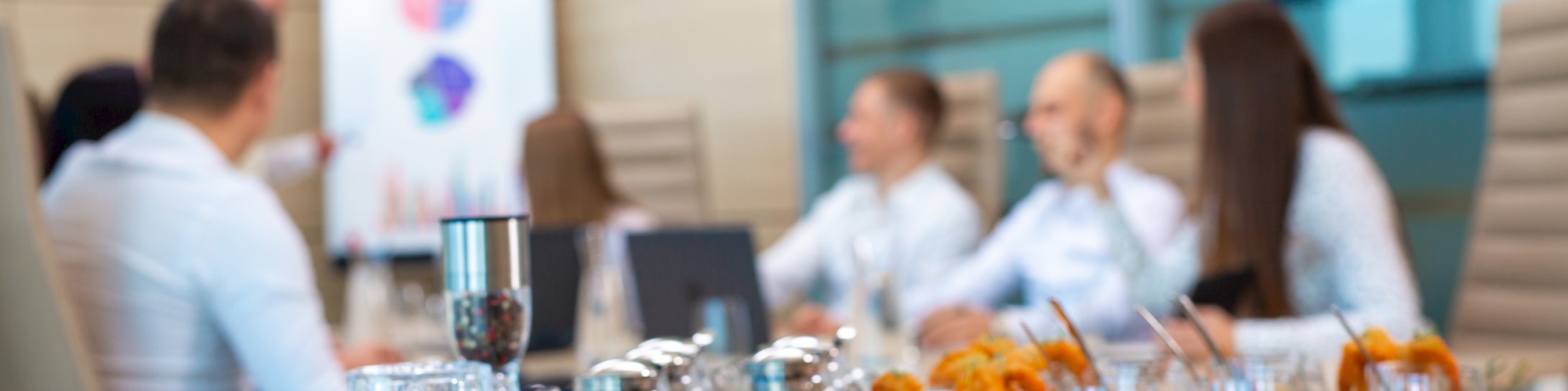 A business meeting is taking place with a diverse array of appetizers and drinks on the table, while a person points at a whiteboard.