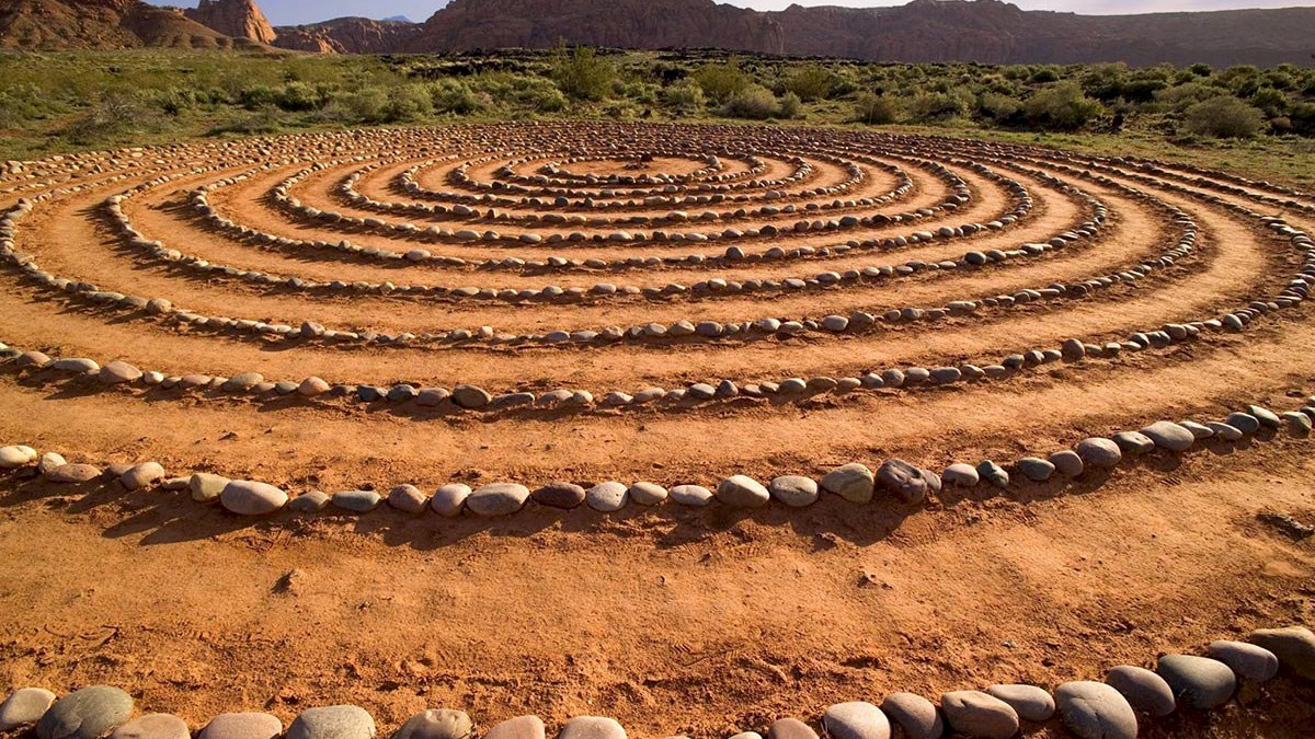 A spiral labyrinth made of stones is set on a sandy ground, with distant hills and greenery in the background under a clear sky.
