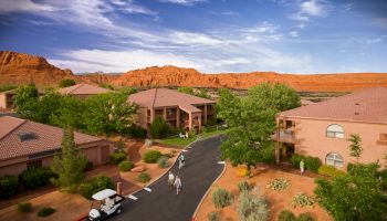 This image shows a scenic resort with several buildings, greenery, people walking, a golf cart, and red-rock mountains in the background under a blue sky.