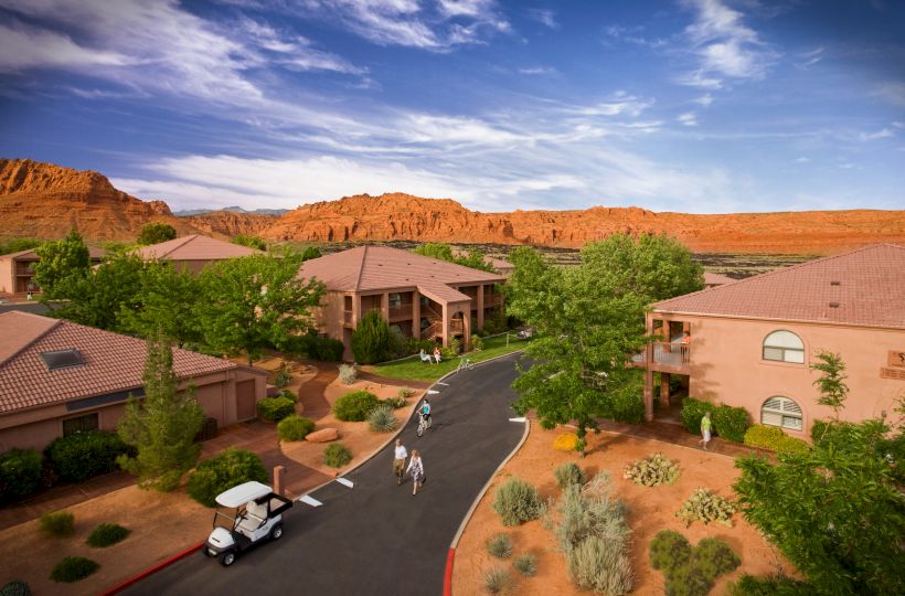 This image shows a scenic resort with several buildings, greenery, people walking, a golf cart, and red-rock mountains in the background under a blue sky.
