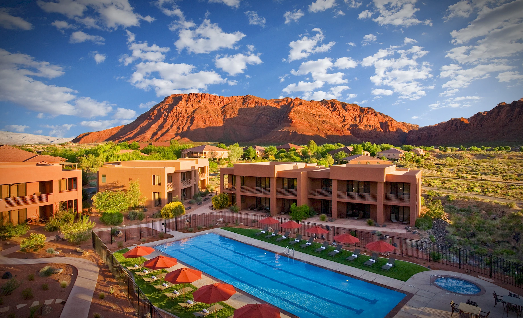 A luxury resort with an outdoor pool, surrounded by desert landscape and red rock formations under a sky with scattered clouds.