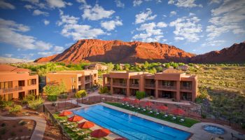 A luxury resort with an outdoor pool, surrounded by desert landscape and red rock formations under a sky with scattered clouds.
