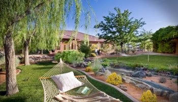A tranquil garden features a hammock, a book, and a pillow under a tree by a stream, with a house and lush greenery in the background.