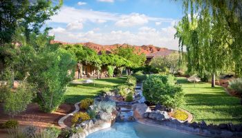 A lush garden featuring a small stream, various trees and plants, a well-maintained lawn, and distant red rock formations under a partly cloudy sky.
