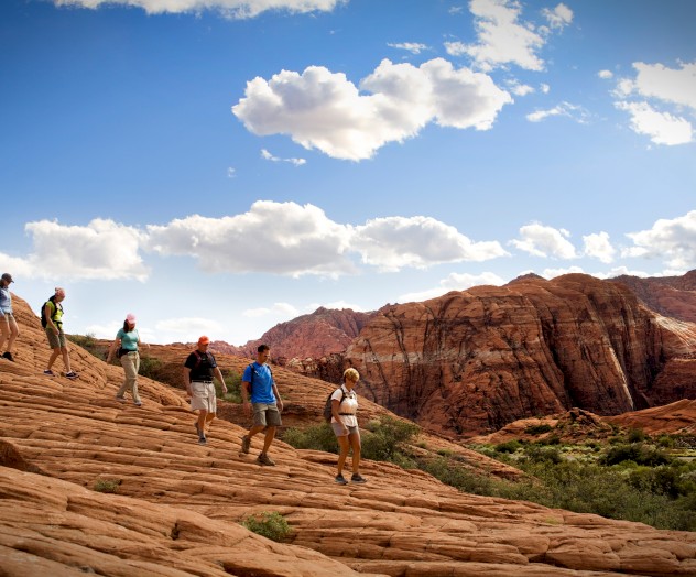 A group of people are hiking on a red rocky terrain under a blue sky with scattered clouds and a backdrop of mountains.
