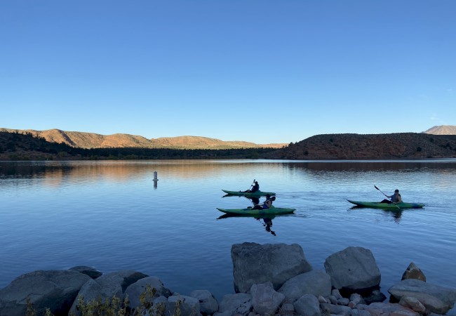 Three kayakers paddle on a calm lake with hilly terrain and clear blue skies in the background.