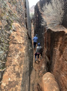 Two people are walking through a narrow, rocky canyon. The walls are steep and covered in different shades of brown and orange rock.