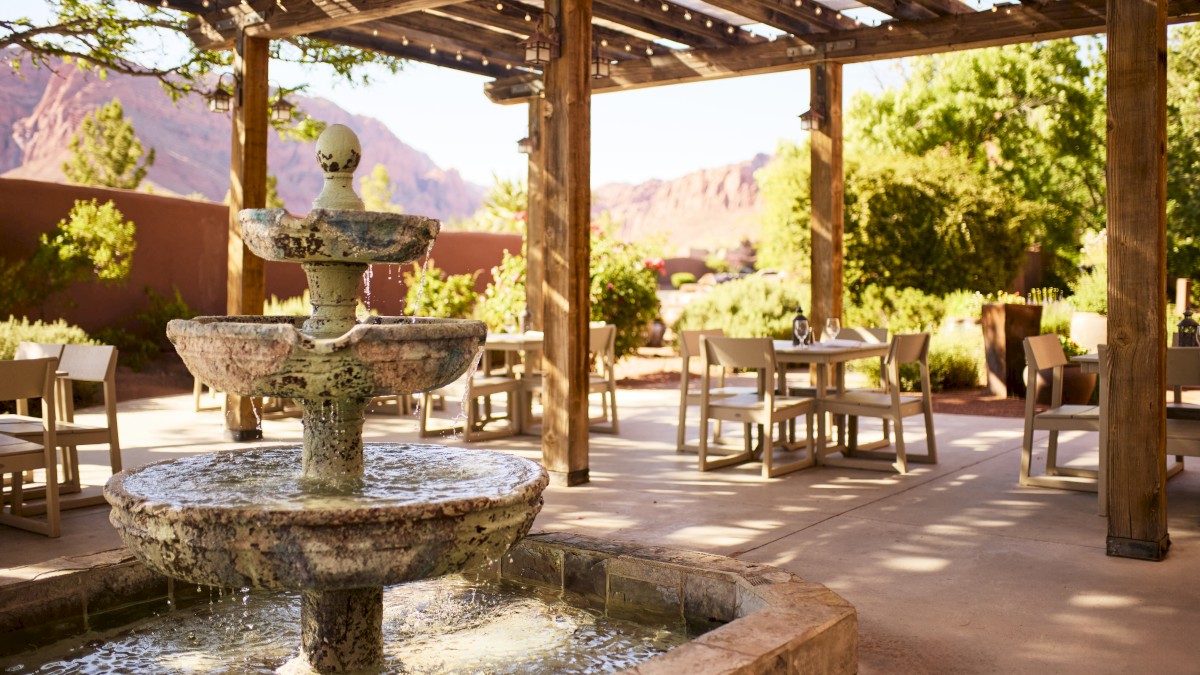 A serene outdoor dining area with a rustic pergola, stone fountain, and wooden tables, surrounded by lush greenery and mountain views.