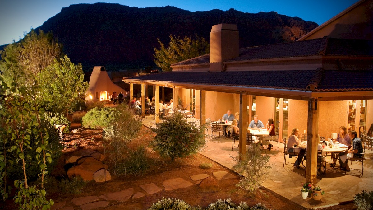 Outdoor dining area at dusk, with people sitting at tables under a covered patio, surrounded by greenery and mountains in the background.