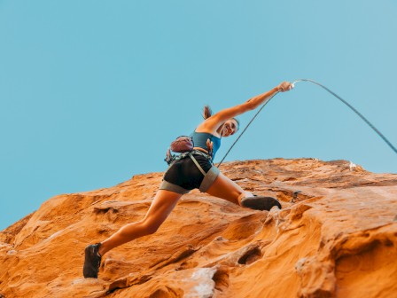A woman climber reaches upward on a red rock face outdoors under a clear blue sky.