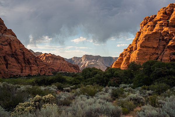 A stunning landscape of red rock mountains, lush greenery, and dramatic cloudy skies, creating a serene and picturesque scene.