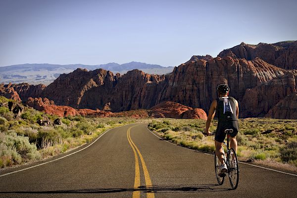 A cyclist rides on a deserted road with red rock mountains and blue sky in the background, wearing a helmet and cycling gear.