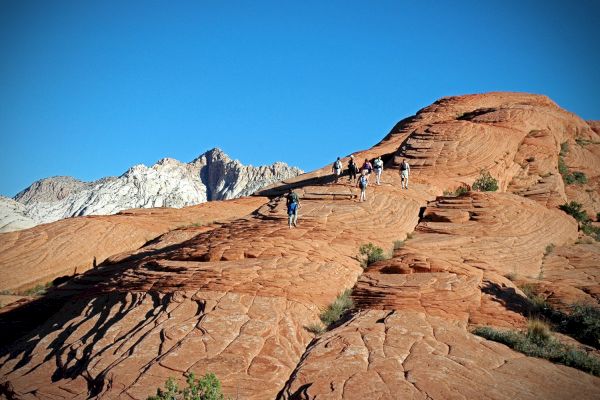 People are hiking on red rock formations under a clear blue sky, with mountains visible in the background.