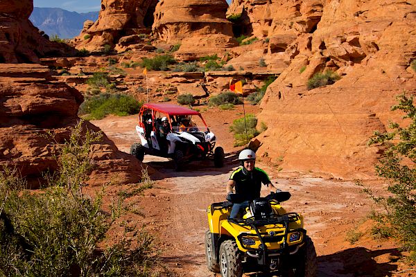 Two off-road vehicles navigate a dirt path amid striking desert rock formations under a clear sky, surrounded by rugged natural terrain.