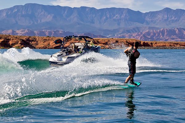 A person is wakeboarding on a lake, being towed by a boat. The scene has mountains in the background under a clear sky.