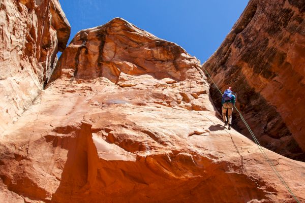 A person is rock climbing on a large orange cliff under a clear blue sky, using climbing gear and ropes for safety, in a canyon-like environment.