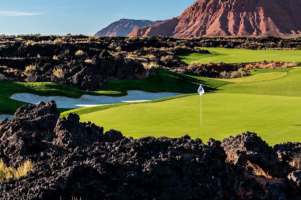 This image shows a golf course with a green field, flagstick, and surrounding rocky terrain with mountains in the background under a clear sky.