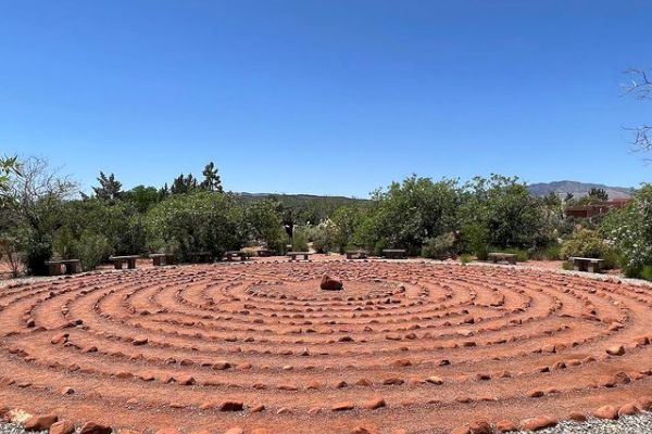 The image shows a large, circular labyrinth made from red rocks, set in a natural outdoor environment with trees and a clear blue sky.