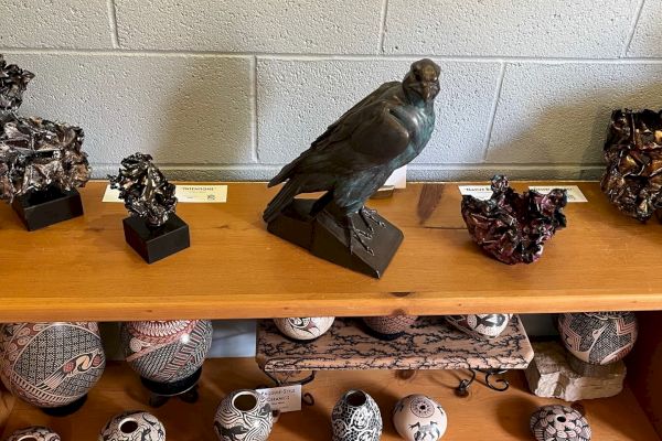 The image shows a display shelf with various artistic objects, including a bird sculpture, pottery pieces, and abstract sculptures, in a room with a gray wall.