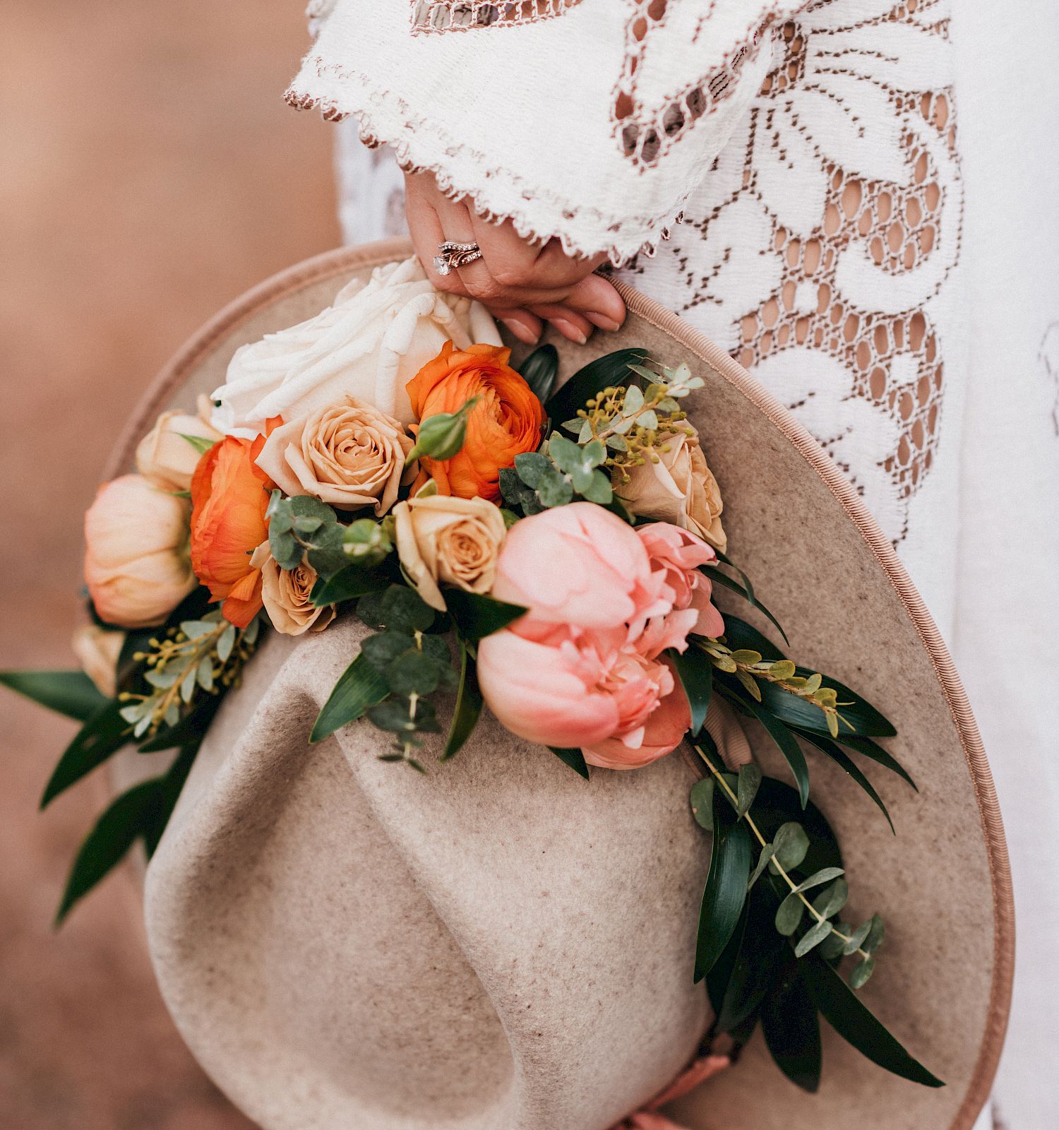 A person in a white, lacy garment holds a hat adorned with vibrant flowers and greenery, creating a rustic yet elegant scene.
