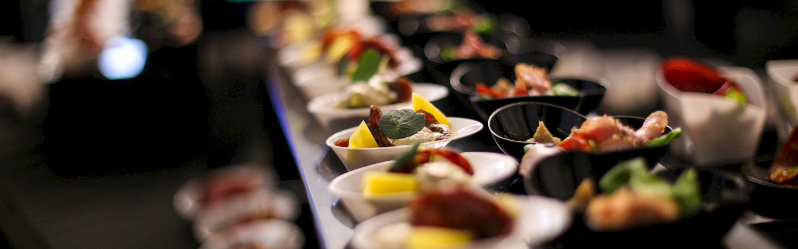 The image shows a display of small, colorful appetizers arranged in bowls and plates, likely at a buffet or catering event, with a blurred background.