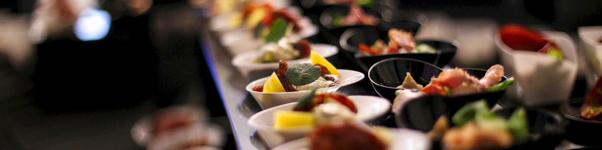 The image shows a display of small, colorful appetizers arranged in bowls and plates, likely at a buffet or catering event, with a blurred background.