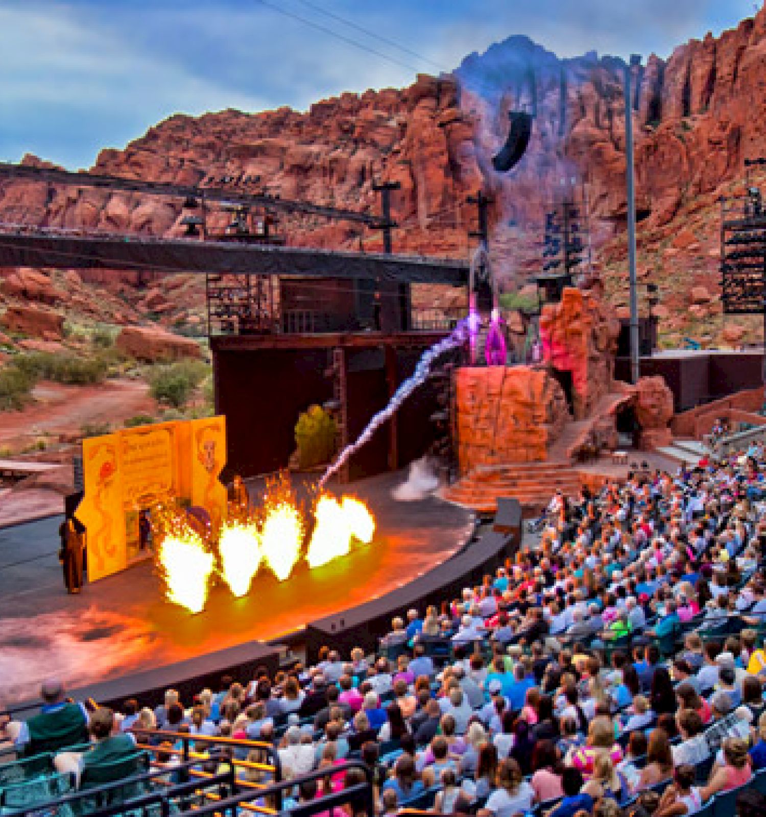 An outdoor amphitheater set in a rocky landscape, filled with people watching a performance featuring fire and special effects.